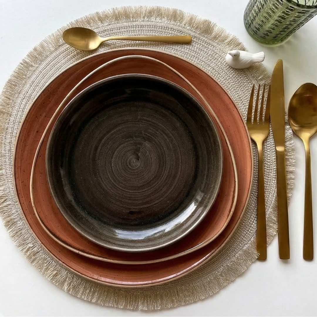 Sottopiatto di tessuto chiaro con bordi frangiati, piatti in ceramica marroni e dorati, posate in oro e un piccolo uccellino bianco.
