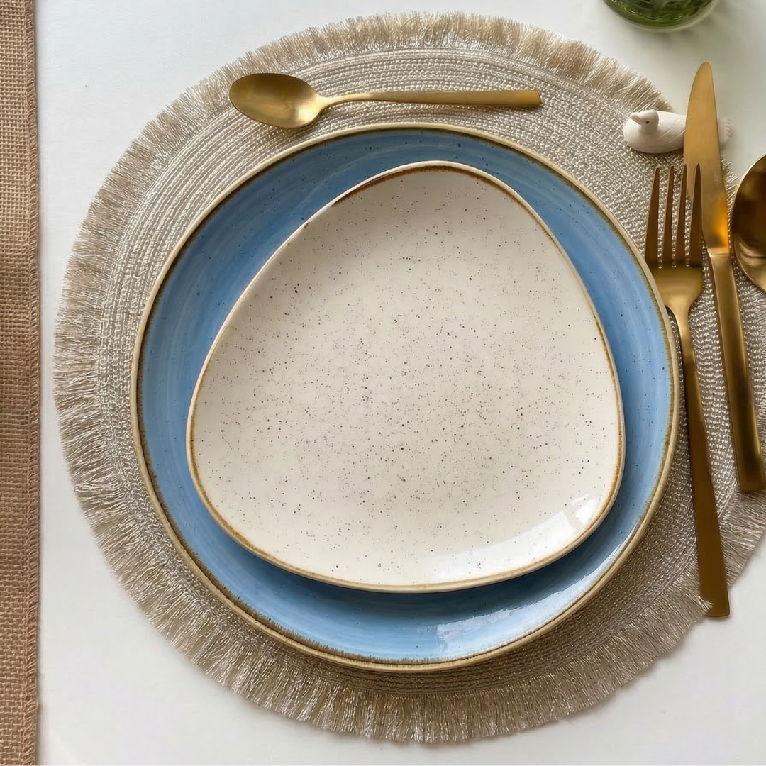 Due piatti sovrapposti su un sottopiatto beige con frange, posate dorate accanto e un oggetto decorativo in cima.