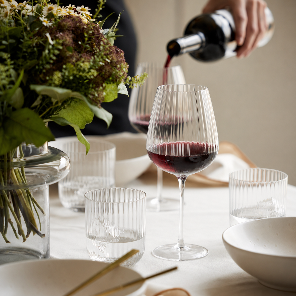 Calice di vino trasparente con vino rosso, bicchieri in vetro e un mazzo di fiori su un tavolo bianco con stoviglie.