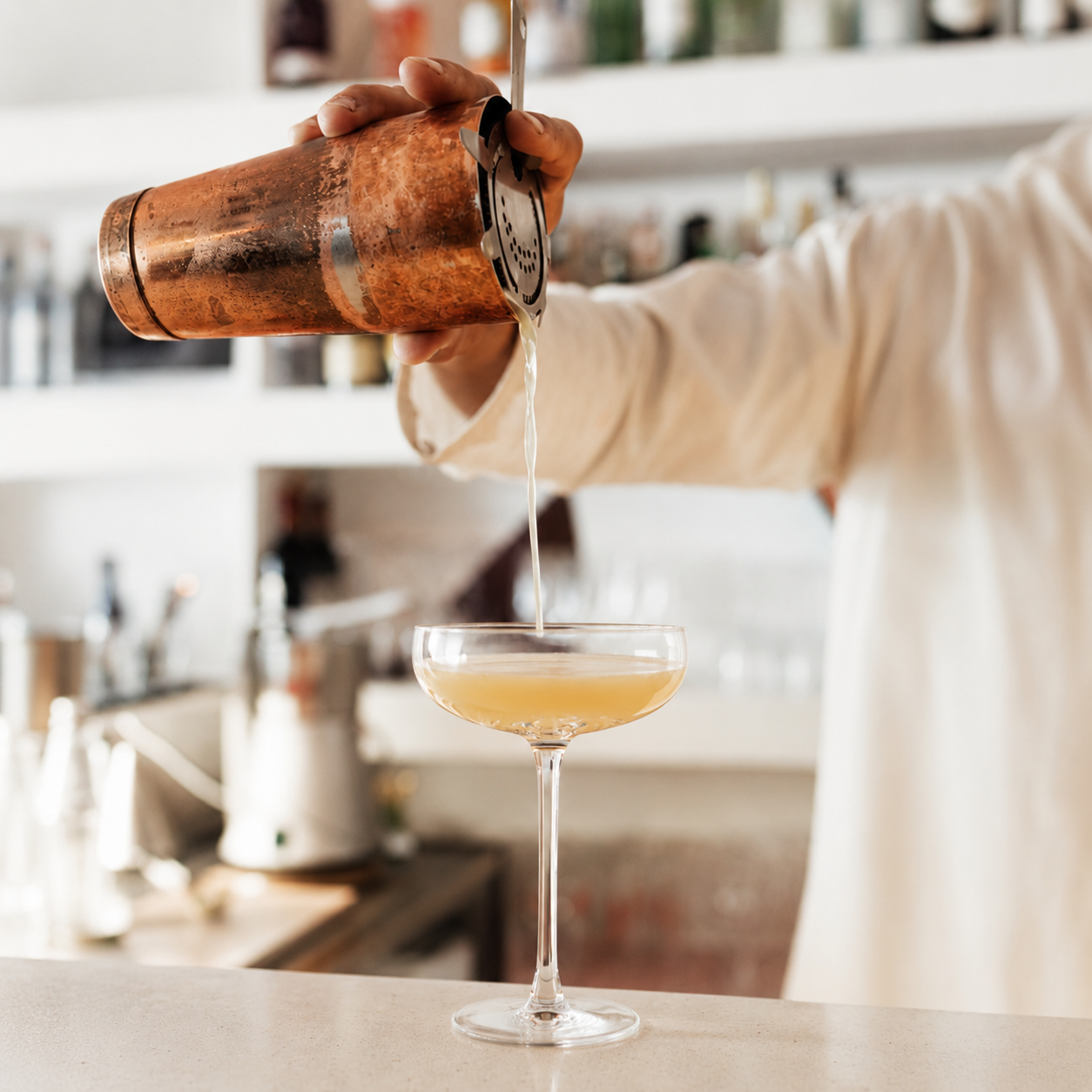 Barman versa un cocktail giallo in una coppa di vetro, mentre tiene un shaker di rame. Sfondo con bottiglie.