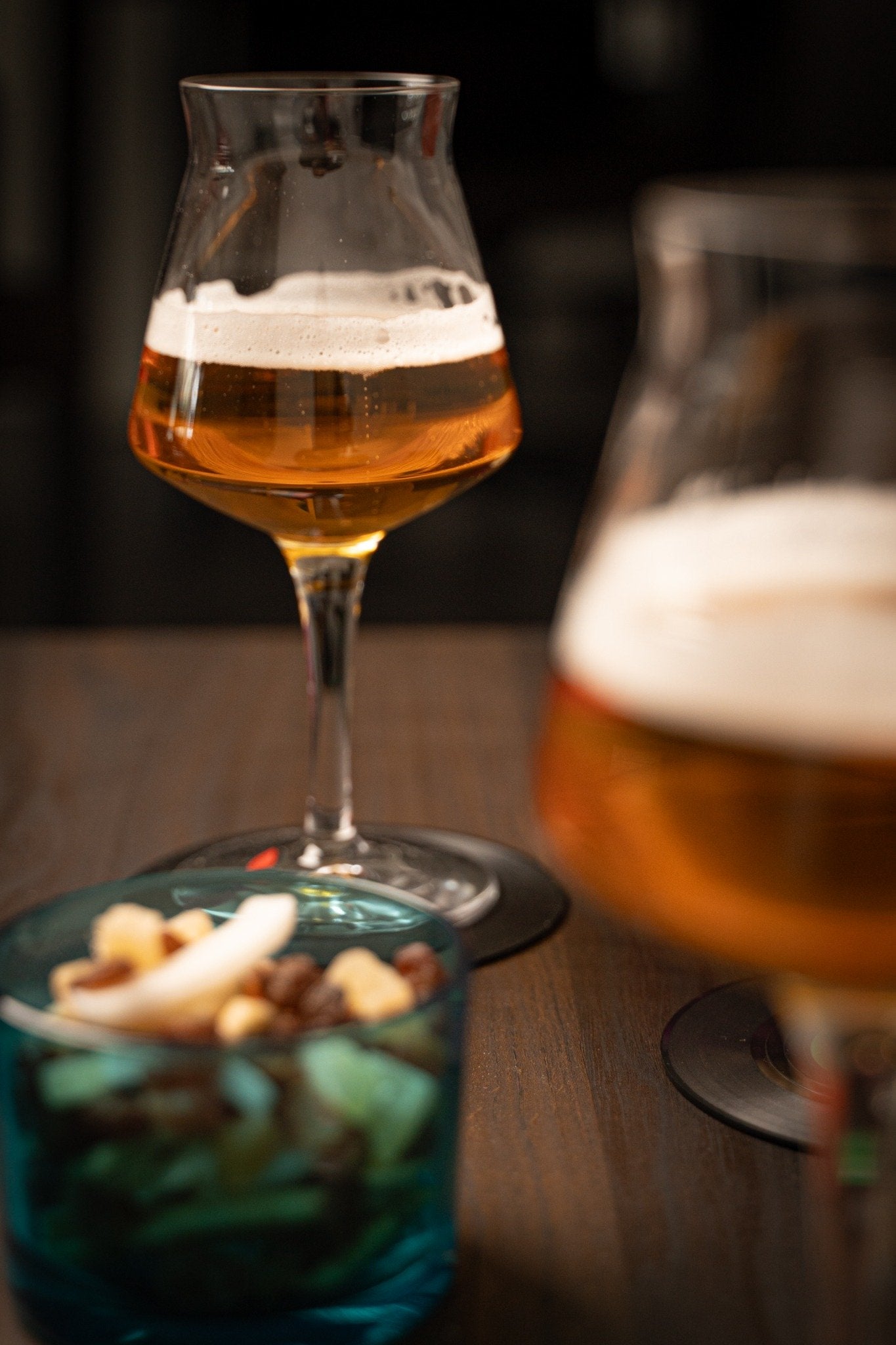 Due calici di birra gialla con schiuma, su un tavolo di legno scuro, accanto a una ciotola di snack misti.