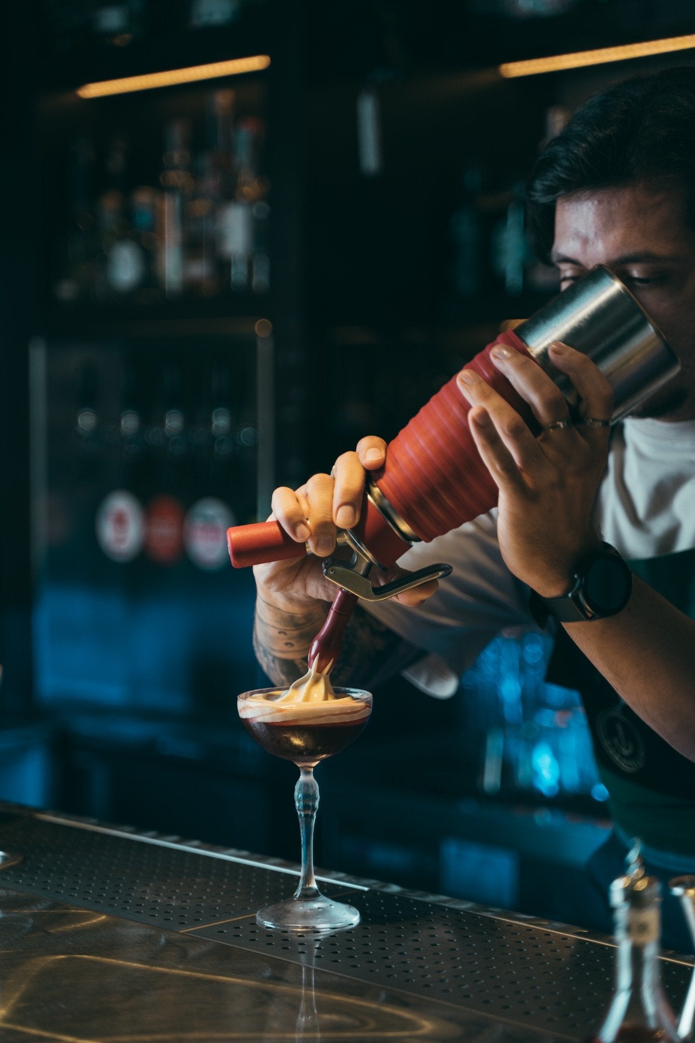 Barman versa una crema su un cocktail scuro in coppetta, mentre sullo sfondo si intravedono bottiglie e luci soffuse.