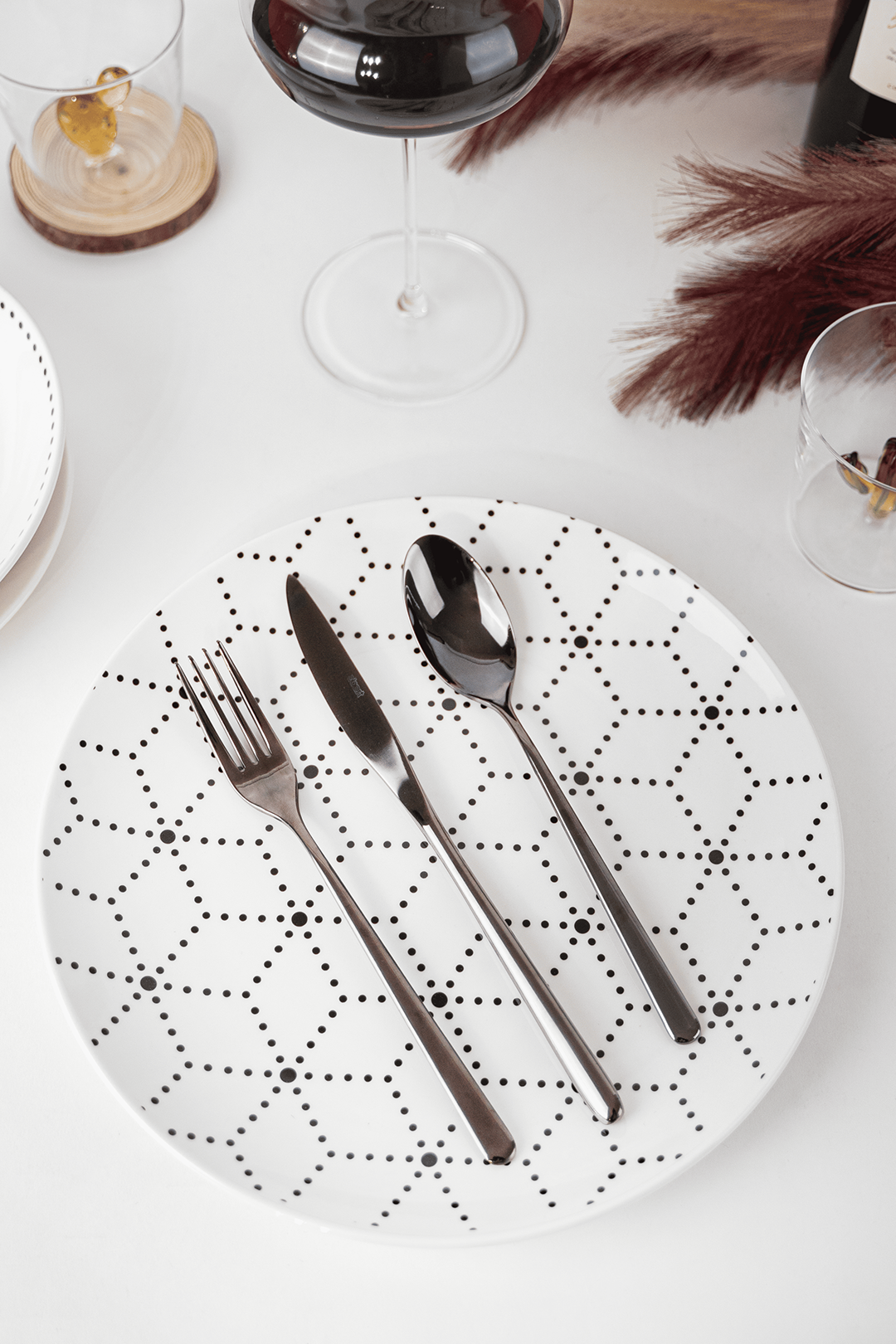 Piatto bianco con motivo geometrico nero, posate in acciaio lucido: forchetta, coltello e cucchiaio, su superficie bianca.