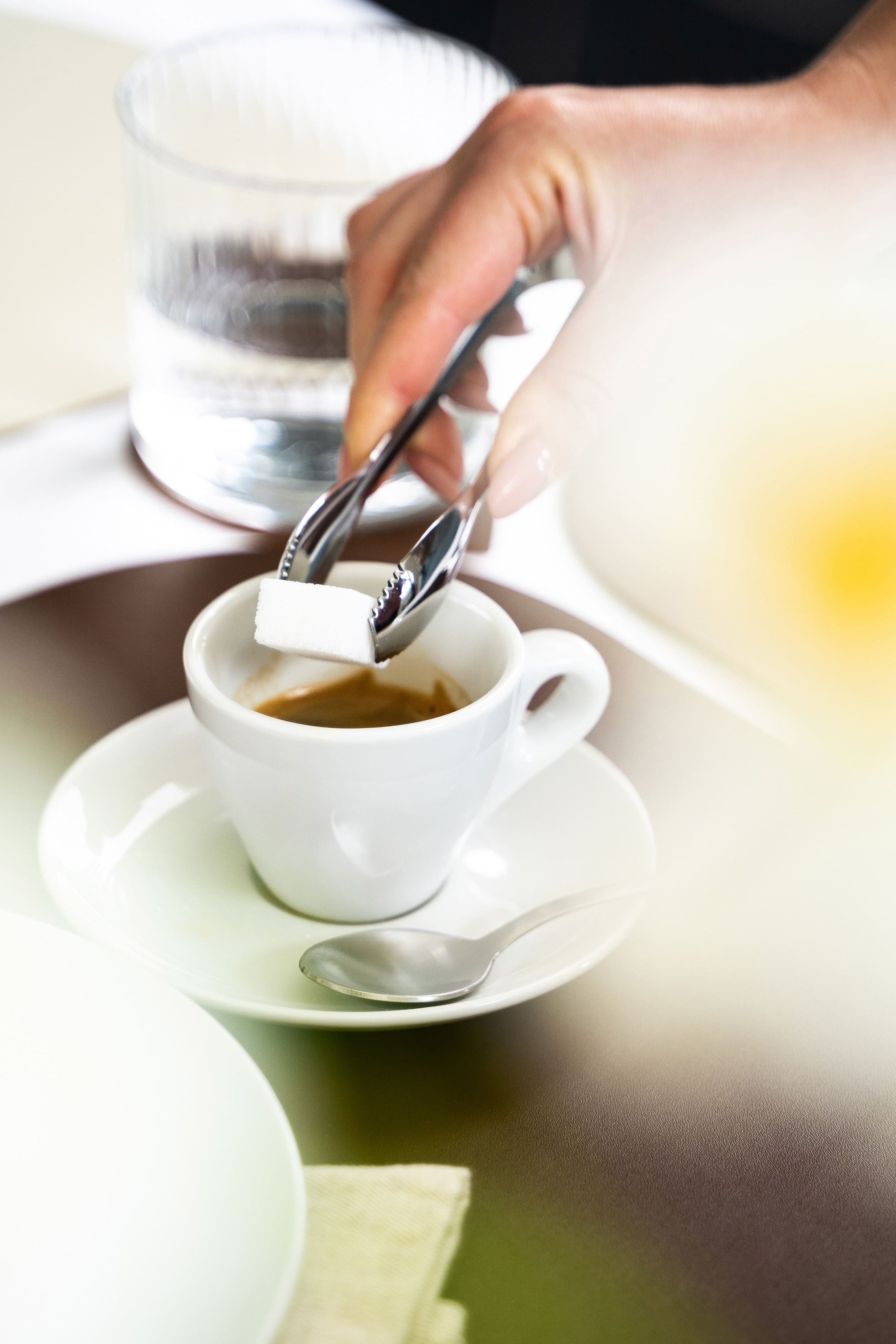Tazza bianca con caffè su piattino, zucchero in pinze metalliche, cucchiaino e vetro d'acqua sul tavolo.