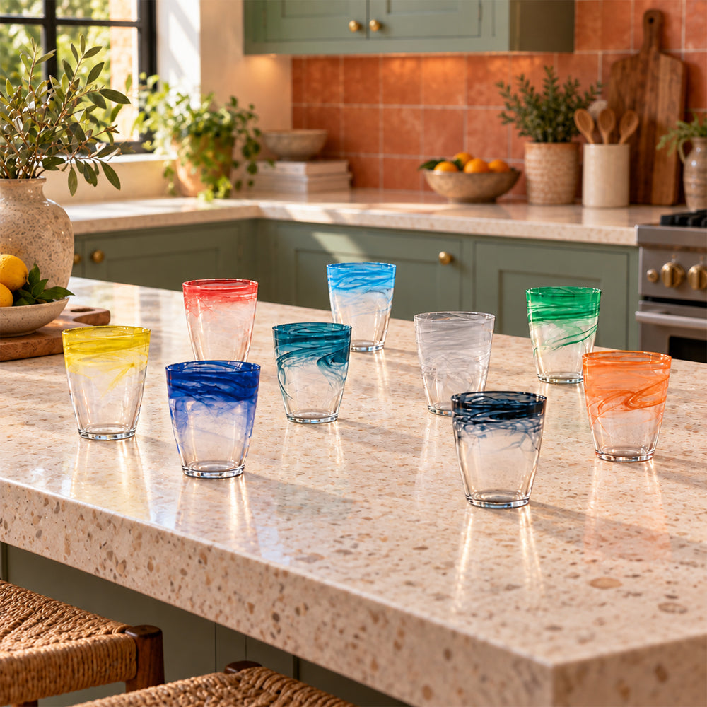 Bicchieri da Acqua sfumati in diversi colori disposti su una tavola apparecchiata, ambientazione soffusa, calda e accogliente.