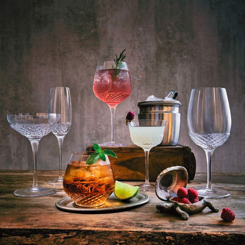 Set di sei bicchieri in vetro trasparente con incisioni, tre cocktail colorati con guarnizioni di frutti e erbe, ghiaccio e lime, su tavolo