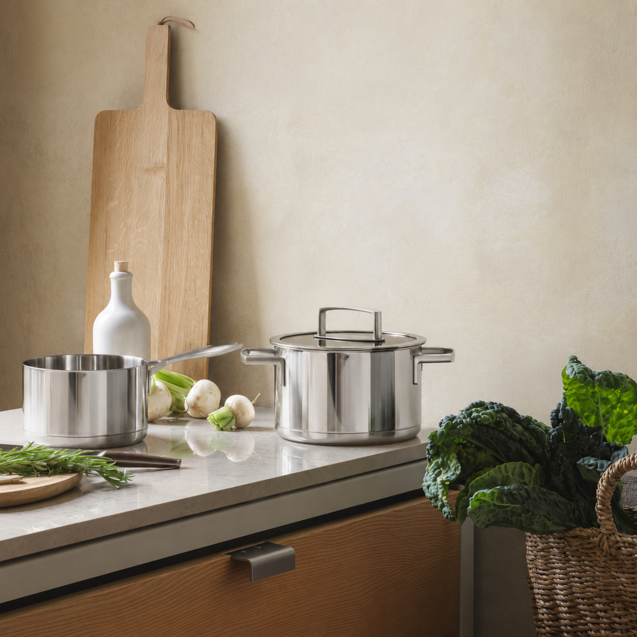 Pentole in acciaio inox su un piano di lavoro. A sinistra, bottiglia bianca e ortaggi freschi. A destra, cestino di verdure. Ambiente cucina.