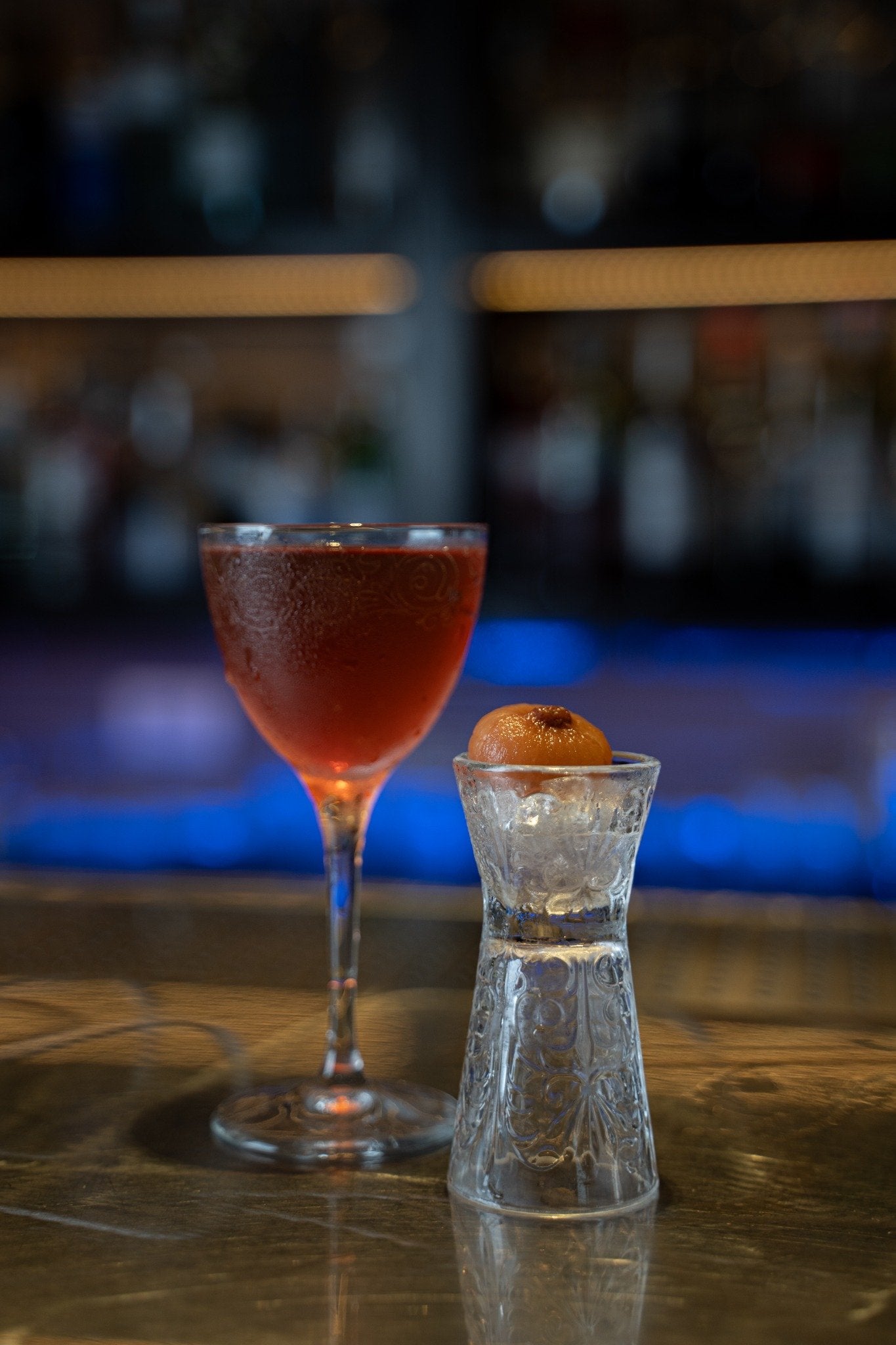 Cocktail rosso in un bicchiere elegante, accanto a un bicchiere trasparente con ghiaccio e una pallina arancione. Sfondo sfocato.
